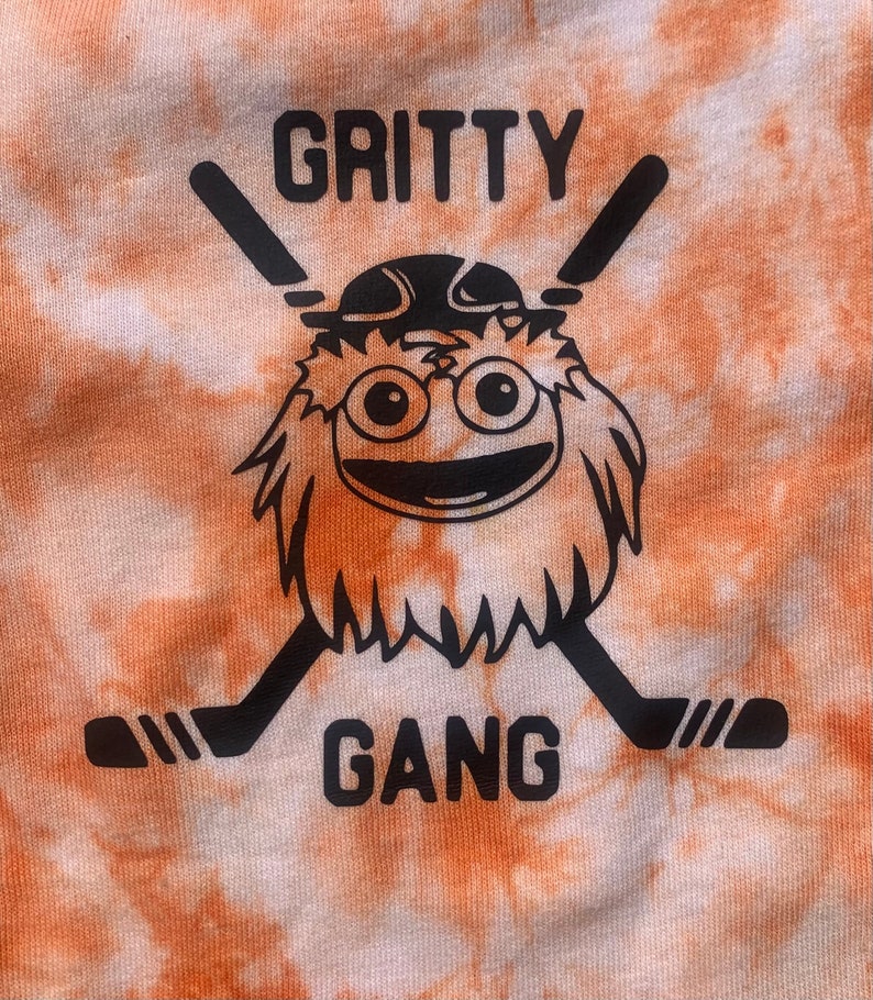 May include: Orange and white tie-dye t-shirt with a black and white graphic of a furry orange creature wearing a hockey helmet and holding two hockey sticks. The text "Gritty Gang" is printed below the creature.