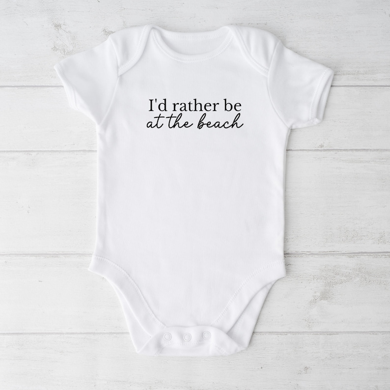 May include: White baby onesie with short sleeves and snaps at the bottom. The onesie has black text that reads "I'd rather be at the beach".