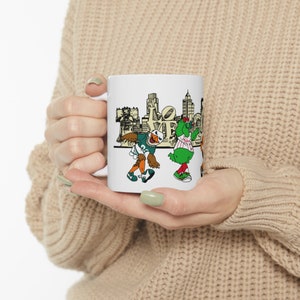 Może przedstawiać: Biały kubek ceramiczny z czarno-białą ilustracją panoramy Filadelfii i maskotek Philadelphia Eagles i Phillies. W ilustracji widoczny jest napis "LOVE".