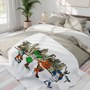 Könnte beinhalten: Eine weiße Decke mit einer bunten Illustration von Maskottchen verschiedener Sportmannschaften aus Philadelphia, darunter die Eagles, Phillies, Flyers und Sixers. Die Maskottchen stehen vor einer Stadtkulisse mit dem Text "BROAD ST" auf einem Straßenschild.