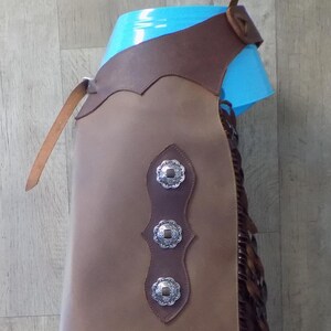 May include: Brown leather chaps with decorative silver conchos and fringe. The chaps have a brown leather belt and are displayed on a blue form. Western wear.