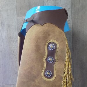 May include: Tan leather chaps with decorative silver conchos and fringe. The chaps have a brown leather belt and are displayed on a blue form. The chaps are a western wear item.