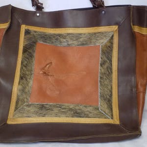 Puede incluir: Un bolso tote de cuero marrón con un gran parche de piel de vaca marrón, tostado y gris en la parte delantera. El bolso tiene una cremallera superior y dos asas.