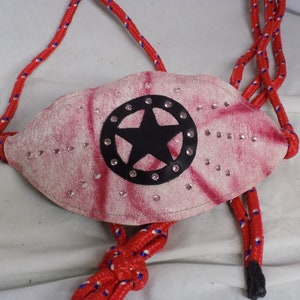 Puede incluir: Un cabestro de cuerda rojo y blanco con una bozal de cuero rosa con una estrella negra en un círculo con diamantes de imitación.