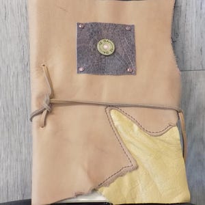 May include: A handmade leather journal with a light brown cover. A metal square with a bullet casing is riveted to the front. The journal is tied with a leather cord. A yellow leather patch is sewn on the front.