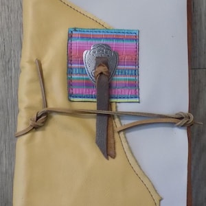 May include: A handmade leather journal with a unique design. The journal features a light yellow cover, a white interior, and a colorful fabric accent with a silver-toned metal clasp. A leather tie secures the journal.