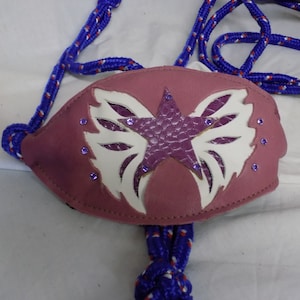 Puede incluir: Un cabezada de cuero rosa con un diseño de estrella blanca y alas. El cabezada tiene diamantes de imitación morados alrededor de los bordes. El cabezada está unido a una cuerda azul y blanca.