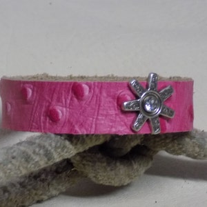 Op de afbeelding: Een roze leren armband met een zilveren sporenbedel. De armband heeft een getextureerd, geëmbost ontwerp.