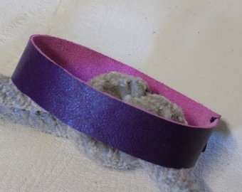 May include: A purple leather bracelet with a pink lining. The bracelet is wrapped around a piece of grey fabric.