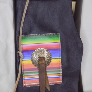 May include: A dark brown leather journal with a colorful striped fabric patch and a metal concho. The journal has a leather strap and a fringe detail.