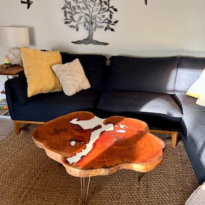 May include: A wooden coffee table with a live edge top and a white epoxy river running through the centre. The table has hairpin legs and is sitting on a woven rug in a living room.