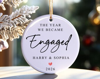 May include: A round white ceramic ornament hanging by a gold string. The text reads THE YEAR WE BECAME Engaged, followed by HARRY & SOPHIA, a small red heart icon, and the year 2026. The surface is smooth and glossy with black lettering.