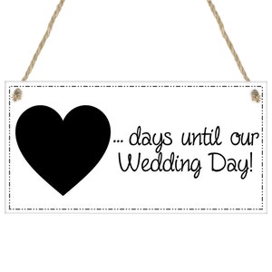 May include: A white rectangular sign featuring a black heart and the text "... days until our Wedding Day!" in black script. It is suspended by a natural-coloured rope.