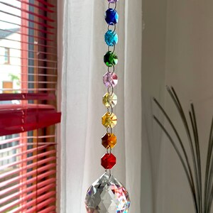 Op de afbeelding: Een regenboogkristallen zonenvanger met een grote, heldere kristal aan de onderkant. De zonenvanger hangt aan een wit gordijn.