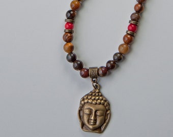 Könnte beinhalten: Halskette mit einem Buddha-Anhänger aus Bronze. Die Kette hat braune und rote Perlen mit bronzefarbenen Abstandshaltern. Der Buddha-Anhänger zeigt ein detailliertes Gesicht mit geschlossenen Augen. Ein spirituelles und dekoratives Accessoire.