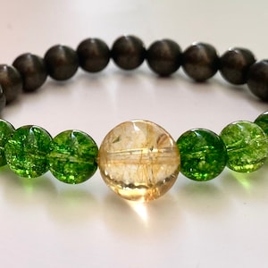 Op de afbeelding: Een armband gemaakt van donkergrijze, groene en gouden kralen. De armband heeft een grote gouden kraal in het midden.
