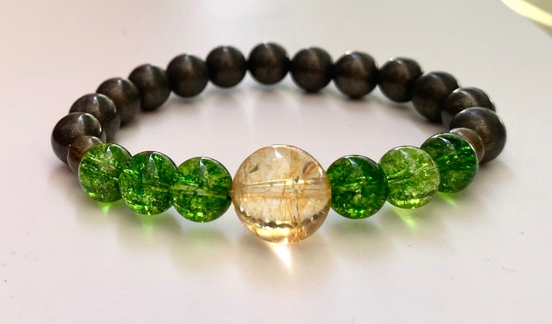 Op de afbeelding: Een armband gemaakt van donkergrijze, groene en gouden kralen. De armband heeft een grote gouden kraal in het midden.