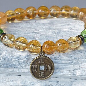 May include: A gold and green beaded bracelet with a gold coin charm featuring Chinese characters. The coin charm has the characters "正道" (Zheng Dao) which means "the right path".