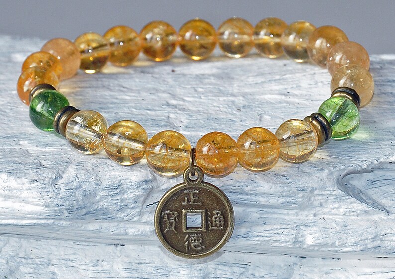 May include: A gold and green beaded bracelet with a gold coin charm featuring Chinese characters. The coin charm has the characters "正道" (Zheng Dao) which means "the right path".
