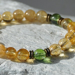 May include: A gold and green beaded bracelet with a gold charm featuring Chinese characters. The bracelet is made with round beads and has a simple design.