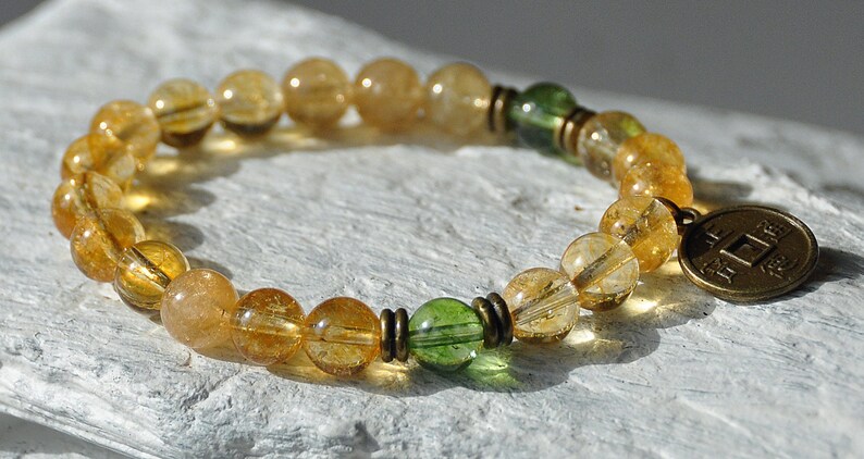 May include: A gold and green beaded bracelet with a gold charm featuring Chinese characters. The bracelet is made with round beads and has a simple design.