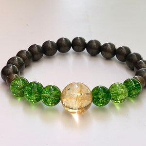 Op de afbeelding: Een armband gemaakt met groene en gouden kralen. De armband heeft een grote gouden kraal in het midden en is omringd door kleinere groene kralen. De armband is gemaakt van een donkergrijs materiaal.