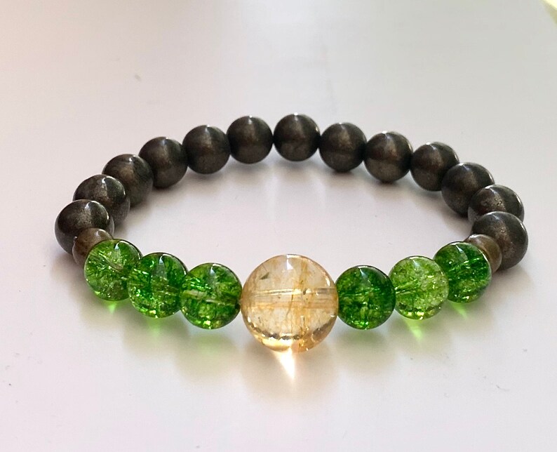Op de afbeelding: Een armband gemaakt met groene en gouden kralen. De armband heeft een grote gouden kraal in het midden en is omringd door kleinere groene kralen. De armband is gemaakt van een donkergrijs materiaal.