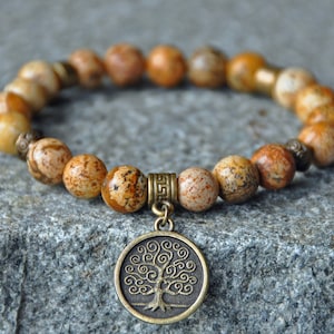 May include: A brown and beige stone beaded bracelet with a bronze tree of life charm. The bracelet is on a grey textured surface.