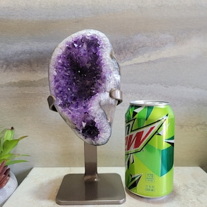 May include: A large amethyst geode with a purple and white crystal formation on a metal stand. The geode is approximately the size of a standard 12-ounce can.