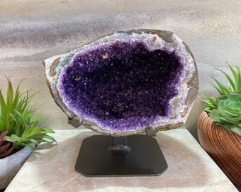 May include: A striking amethyst geode, displaying a rich purple crystal interior. The geode is presented on a black metal stand, with a rugged, natural exterior. Decorative potted plants are positioned on either side of the geode.