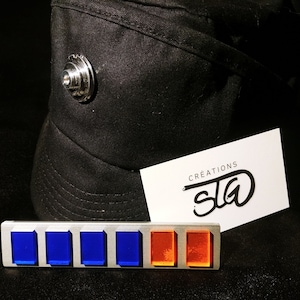 Puede incluir: Una insignia de metal plateado con secciones rectangulares azules y naranjas. La insignia está sobre una superficie negra con un sombrero negro y una tarjeta blanca con el texto "CREATIONS  S/G" en ella.