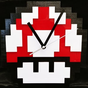 Puede incluir: Un reloj negro, blanco y rojo con forma de seta Super Mario. Las manecillas del reloj son negras y plateadas.