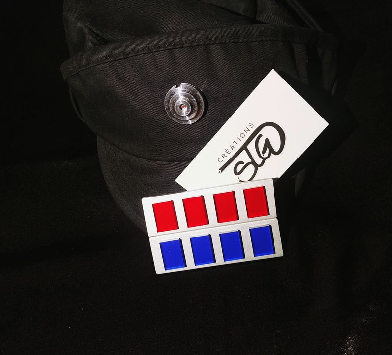 May include: A black cap with a silver pin and a white card with the text "SIG CREATIONS". A silver metal patch with six red and blue squares.