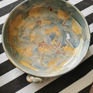 May include: A large, hand-painted ceramic bowl with a wide rim and two rope-style handles. The interior features a swirling design in shades of blue, yellow, and brown. The bowl is set against a black and white striped background.