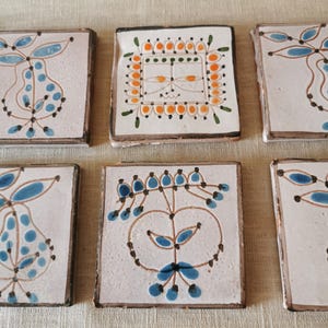 May include: A collection of six square ceramic coasters with hand-painted designs. Each coaster features a white background with brown edges and a unique pattern in blue, orange, and brown. The designs include floral and fruit motifs.