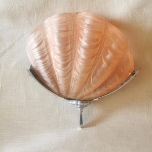 May include: A wall sconce featuring a frosted, peach-colored glass shade shaped like a large seashell with ribbed textures. The shade is supported by a polished silver-toned metal frame that includes a central decorative finial at the base.