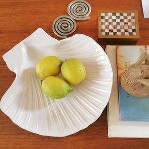 Peut inclure: Un bol en céramique blanche en forme de coquillage contenant trois citrons jaunes, accompagné de deux objets métalliques en spirale argentés, une petite boîte rectangulaire à damier et une figurine sculptée beige posée sur un livre bleu et blanc.