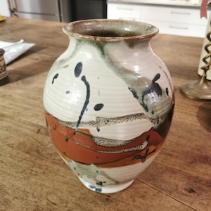 Peut inclure: Un vase en céramique avec un corps arrondi et un bord évasé. La surface présente une base de couleur crème avec des stries horizontales, accentuée par des éclaboussures abstraites de brun foncé, de noir et une large bande orange terre cuite.