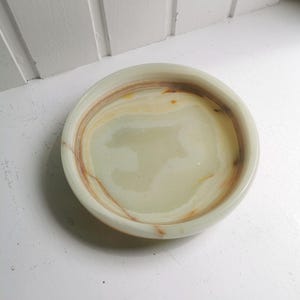 Könnte beinhalten: Eine runde, flache Schale aus poliertem Stein mit glatter, durchscheinender Optik. Die Schale hat eine blass-minzgrüne Grundfarbe mit wirbelnden, organischen Bändern aus warmen bernsteinfarbenen und braunen Maserungen im Inneren und am Rand.
