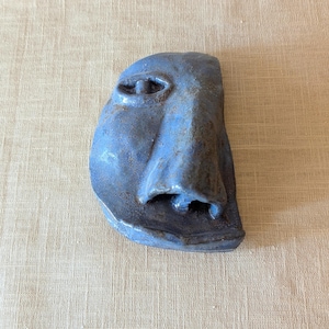 Könnte beinhalten: Eine blaue Keramik-Gesichtsskulptur mit strukturierter Oberfläche. Die Skulptur zeigt eine Nase, ein Auge und einen Teil des Gesichts. Das Kunstwerk ist ein einzigartiges Dekorationsobjekt.