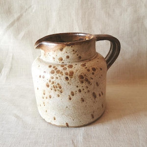 Może przedstawiać: Ceramiczny dzbanek z nakrapianą kremowo-brązową glazurą. Naczynie posiada zaokrąglony korpus, krótką szyjkę, szeroki dzióbek oraz solidny, zakrzywiony uchwyt. Powierzchnia wykazuje wzór ciemnobrązowych plamek na jasnym tle o nakrapianej fakturze.
