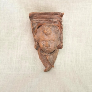 May include: A brown terracotta wall corbel featuring a sculpted human face at the center. The architectural element has a rectangular top section and a tapered base, with a textured, weathered finish across the entire surface of the clay sculpture.