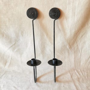 Pode incluir: Um par de castiçais de parede em metal preto com uma haste vertical longa rematada por um design em espiral circular e um prato circular para gotas com um encaixe cilíndrico central para segurar uma vela cónica.