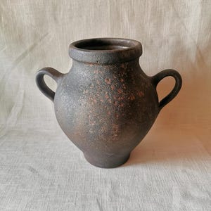 Może przedstawiać: Ciemnobrązowe, teksturowane naczynie ceramiczne o szerokim, zaokrąglonym korpusie i dwóch symetrycznych uchwytach po bokach. Powierzchnia ma cętkowany, zwietrzały wygląd z czerwonobrązowymi odcieniami oraz gruby, wyraźnie zaznaczony brzeg otworu.