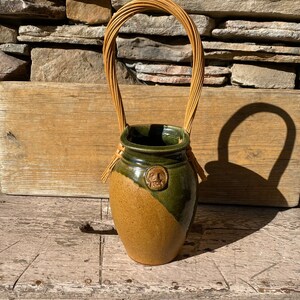 May include: A handcrafted ceramic vase with a woven handle. The vase features a two-tone design with a tan and green color scheme. A small face is embossed on the green section. The handle is made of woven twigs.