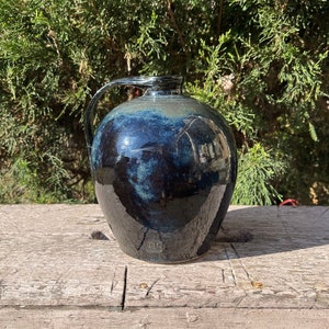 May include: A dark blue ceramic pitcher with a handle. The pitcher has a glossy finish and is sitting on a wooden surface.