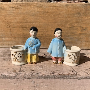 May include: Two ceramic figurines of Asian men in blue and yellow robes, each holding a small white planter with gold floral designs.