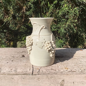 May include: White ceramic vase with a grapevine design. The vase has a textured surface and is decorated with raised grapes.