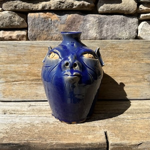 May include: A blue ceramic vase shaped like a face with large eyes and a wide mouth. The vase has a textured surface and a glossy finish.