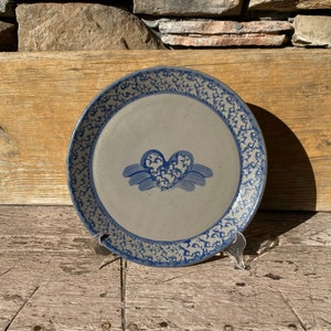 May include: A blue and white ceramic plate with a heart design. The heart is surrounded by a blue and white floral pattern. The plate is on a wooden surface.